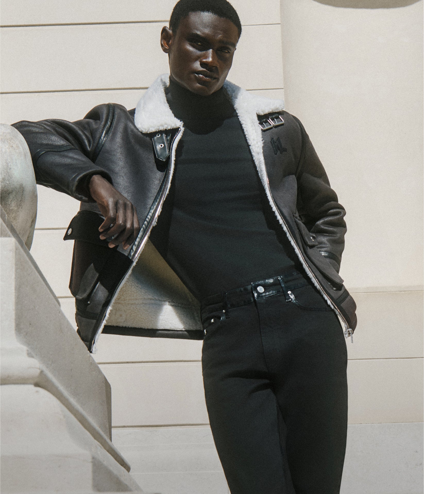Man wearing a gray shearling jacket with a black turtleneck and pants on a white background