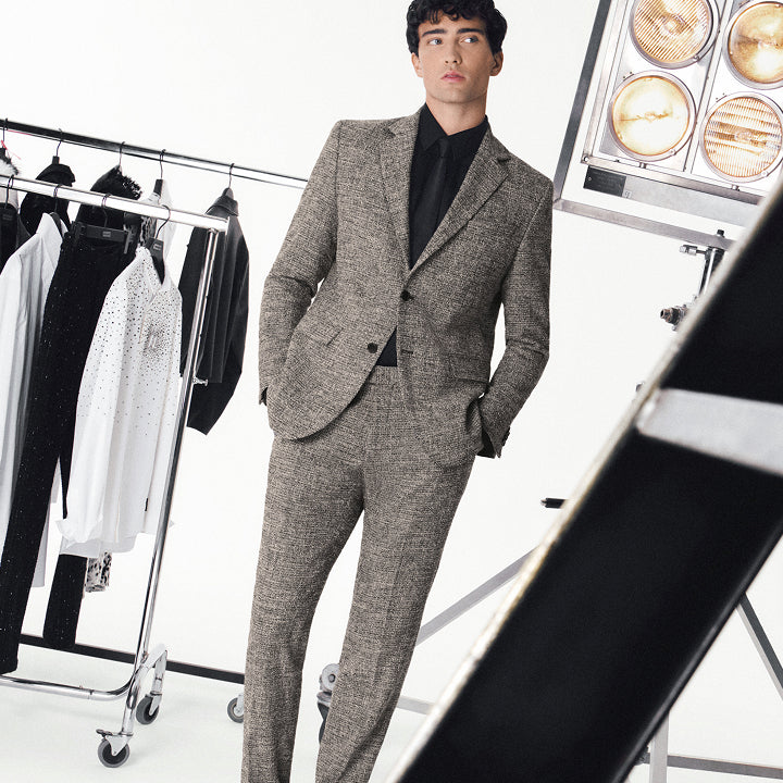Man wearing a gray suit standing in a clothing store with racks of clothes and lighting equipment.