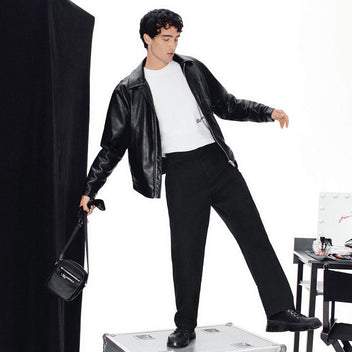 Man in black leather jacket and pants standing on a silver suitcase with a white background