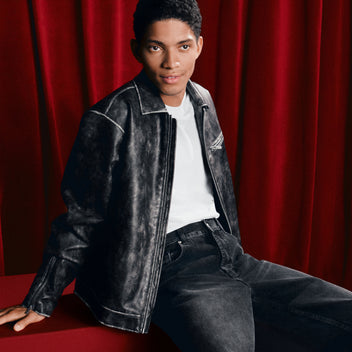 Person wearing a black leather jacket and jeans sitting against a red curtain.