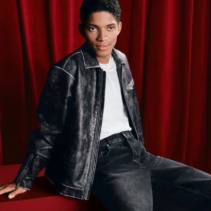 Person wearing a black leather jacket and jeans sitting against a red curtain.