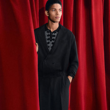 Person wearing a black outfit with a patterned shirt against a red curtain background