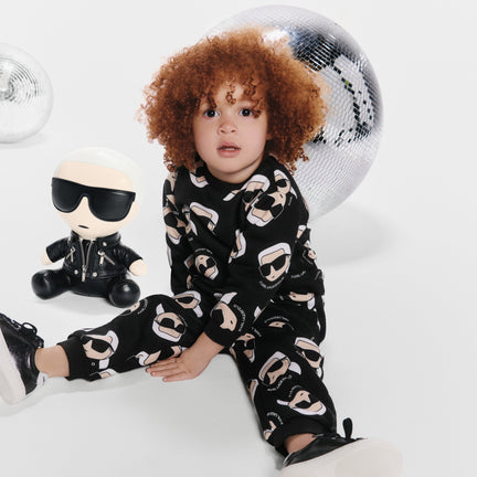 Child wearing a black outfit with white patterns, sitting on a white surface with a toy and disco ball in the background.
