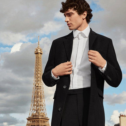 Man in a suit standing in front of the Eiffel Tower with a cloudy sky.