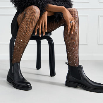 Person wearing black Chelsea boots and fishnet stockings sitting on a black stool.