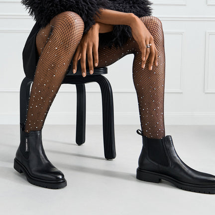 Person wearing black Chelsea boots and fishnet stockings sitting on a black stool.