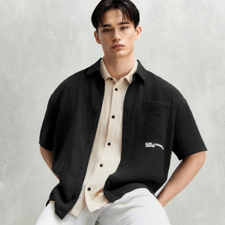 Man wearing a black shirt over a cream shirt with a visible brand logo on a light gray background