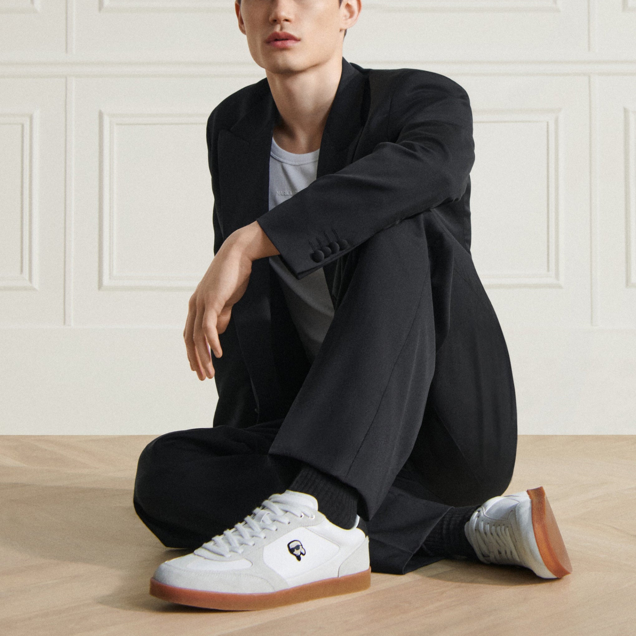 Man wearing a black coat and white sneakers sitting on a wooden floor.