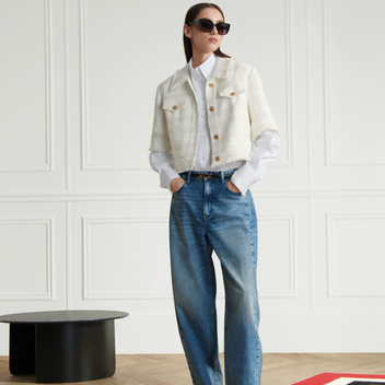 Woman wearing a white jacket, white shirt, blue jeans, and black shoes in a minimalistic room.